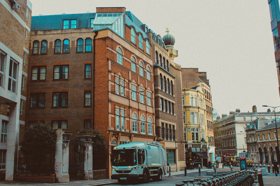 The image shows a city street with multi-storey historic buildings featuring brick facades, large windows, and decorative architectural details. A Westminster Movers van is parked on the pavement near