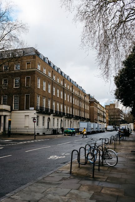 The image shows a city street with multi-storey historic buildings featuring brick facades, large windows, and decorative architectural details. A Westminster Movers van is parked on the pavement near