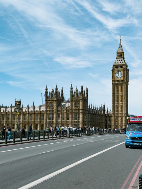 A street scene in Westminster, London, featuring the iconic Big Ben clock tower attached to the Houses of Parliament, with a line of pedestrians walking along the pavement. In the foreground, a parkin